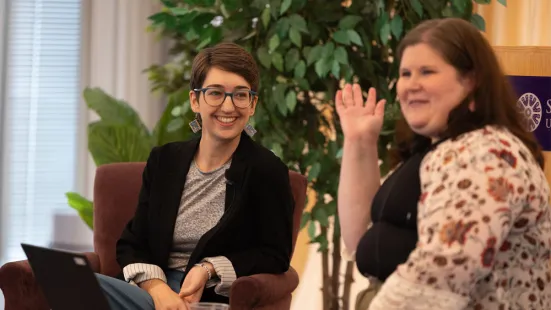 Left to right: A woman with short brown hair, glasses, and a black blazer over a gray shirt smiles at a woman with medium-length brown hair and a floral cardigan over a black shirt, who is speaking.