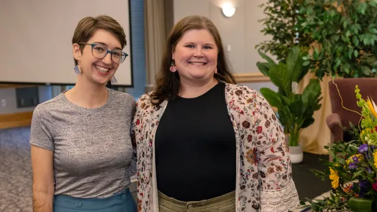 Left to right: a woman with short brown hair, glasses, and a gray shirt standing next to a woman with medium-length brown hair and a floral cardigan over a black shirt. Both women are smiling.