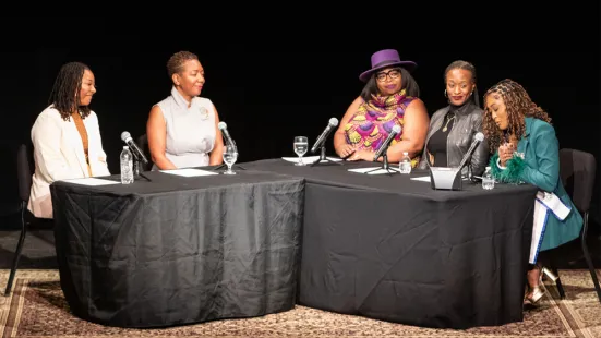 A group of panelists sitting together on a stage at a table with black tablecloth and microphones.