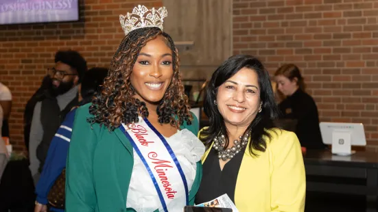 Two women standing together and smiling at the camera. The woman on the left is wearing a crown and sash.