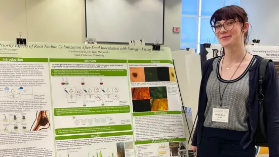 A photo of Caroline Davis in front of a green poster presenting scientific research and data. She is wearing glasses, a striped black and white shirt, a navy blue cardigan, navy blue pants, a necklace, a black purse, and a lanyard with her nametag. Her hair is tied into a ponytail. She is smiling.