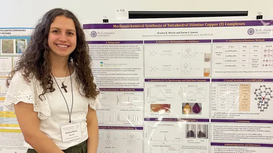 Caroline Davis in front of a purple and white poster presenting scientific research and data. She is smiling and wearing a white blouse with green pants, a cross necklace, and a lanyard with her nametag. Her hair is long and curly.
