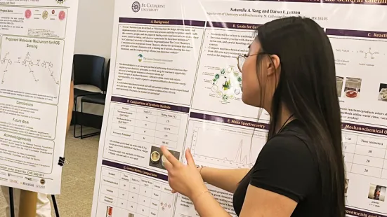 A side-profile of Naturelle Vang presenting research on a purple and white poster. She is pointing to an image while talking. She has long black hair, glasses, and is wearing a black shirt.