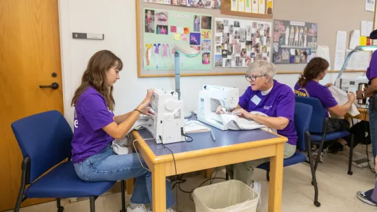 Students and a faculty member using sewing machines in a classroom decorated with fashion sketches.