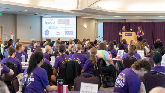 Groups of people wearing purple shirts sit at tables in front of a podium and a screen.