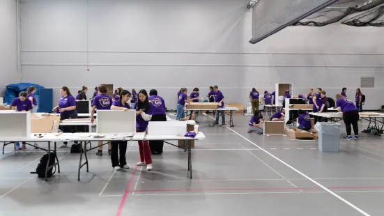 In a gray gym, people in purple shirts work at tables, assembling boxes.