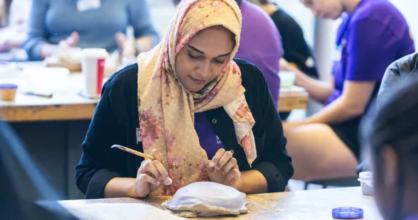 A woman holding a paintbrush works with a mound of clay.