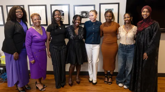 A group of eight women dressed in formal clothes stand together and smile.