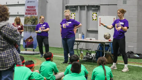 Staff and students gesture as they teach a group of kids sitting on the ground watching.