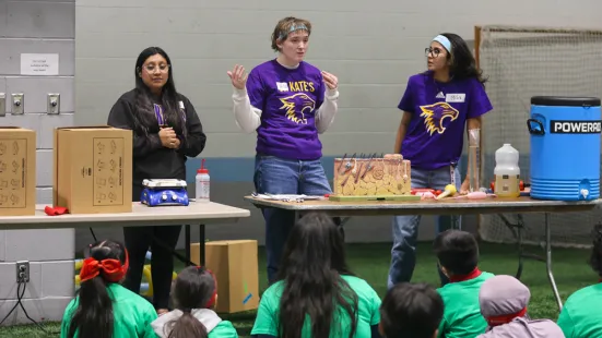 Students stand behind a table and teach a group of children sitting down.