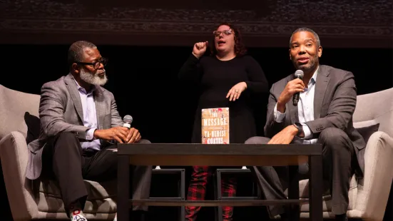 Duchesne Drew and Ta-Nehisi Coates sit in armchairs on stage; in between them is a table with a book standing on it.