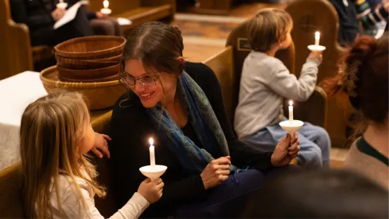 A woman sitting a pew smiles at a child holding a candle.