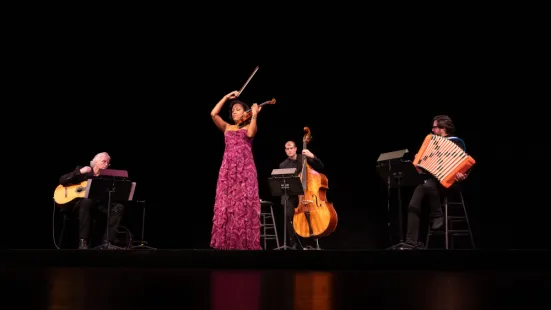 Kelly Hall-Tompkins plays violin onstage, surrounded by a guitarist, bassist, and accordionist