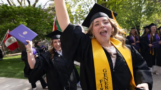 A graduate in a cap and gown waves her arm in the air and smiles.