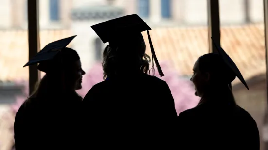 Three graduates in caps and gowns are silhouetted against a window.