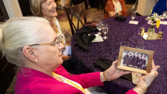 A woman with white hair and a pink jacket holds a photo of graduates in caps and gowns.