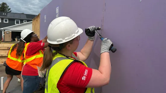 A person wearing a neon safety vest and a hard hat hammers a nail into a purple wall.