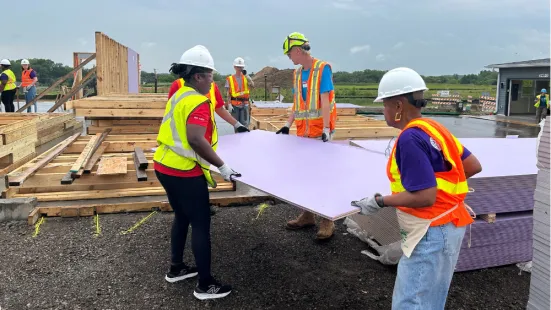 Volunteers wearing neon safety vests and hard hats carry a purple panel of drywall.