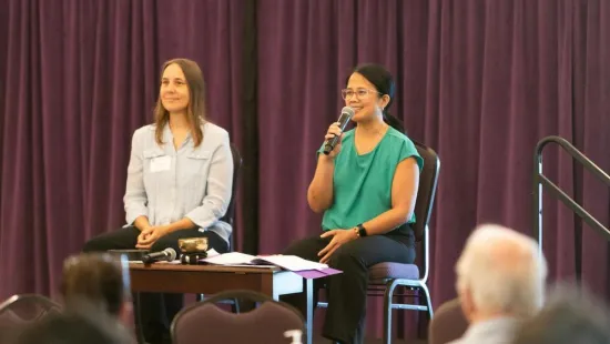 Two women sit on a stage, and one speaks into a microphone.