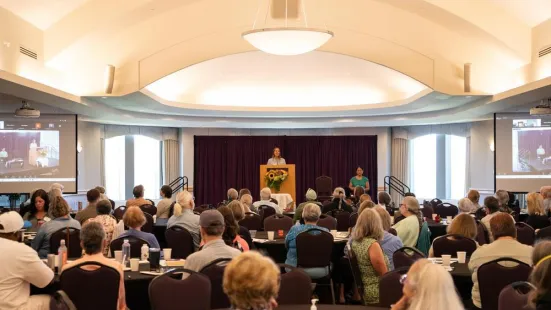 Someone stands at a podium; in front of them is an audience of people sitting at tables.