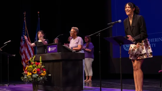 A student stands on stage in front of a microphone; other speakers stand in the back.