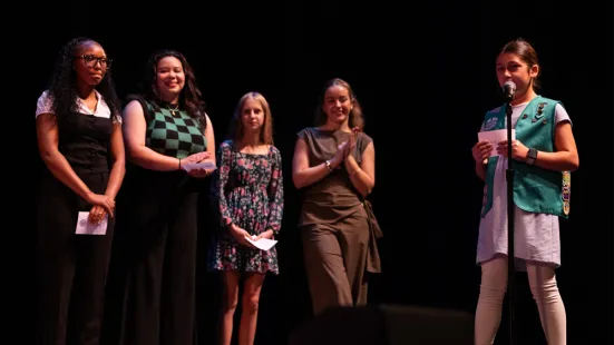A young girl in a Girl Scout vest stands at a microphone on stage; other girls are lined up to the side.