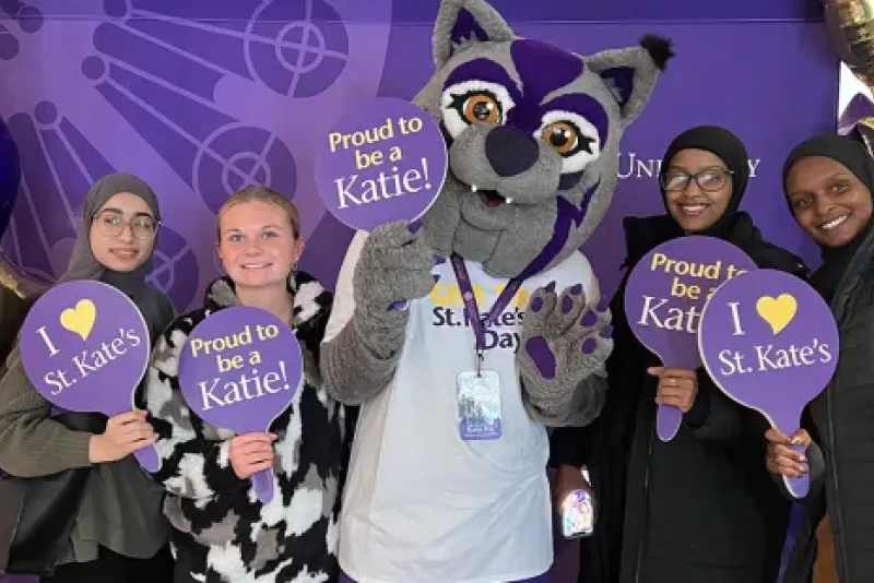 alt="Students hold quote 'I love St. Kate's' and 'Proud to be a Katie' signs while smiling and posing with Katie Cat in front of a purple background.