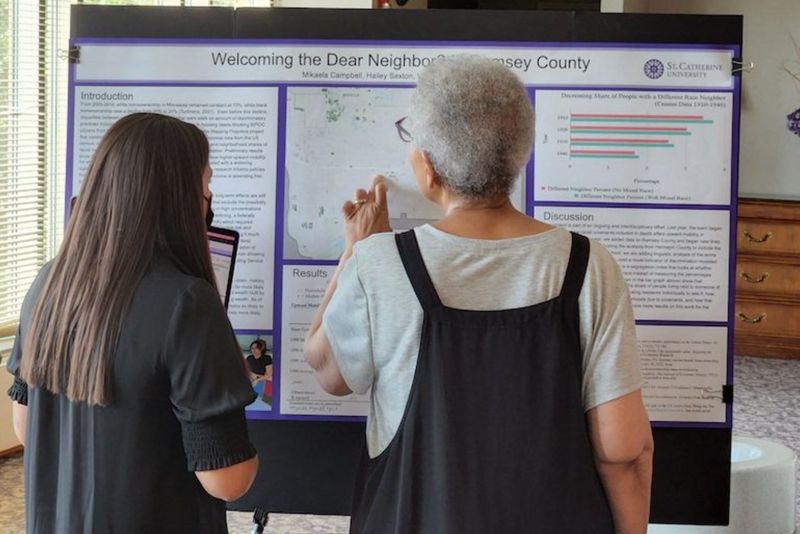 Two people, with their backs to the camera, look closely at a poster that reads "Welcoming the Dear Neighbor? Ramsey County"