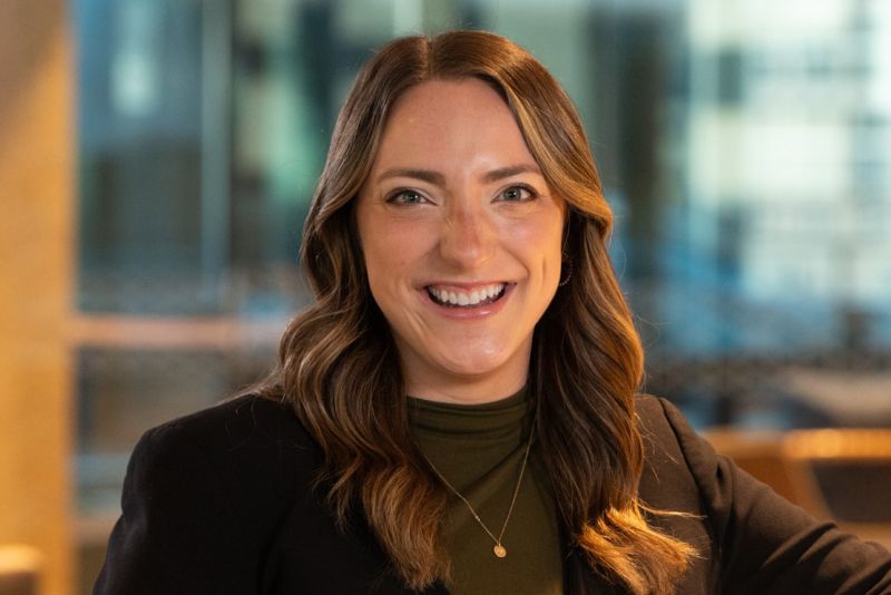A portrait of Maura Bremer Peterson. She is smiling and wearing an olive green turtleneck, brown blazer, and a gold necklace. Her hair is down.