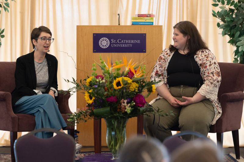 Two women sit in armchairs talking. The woman on the left has short brown hair, glasses, a black blazer over a gray shirt, and turquoise pants. The woman on the right has medium-length brown hair, a floral cardigan over a black shirt, and olive green pants. Between them is a vase of flowers and a podium.