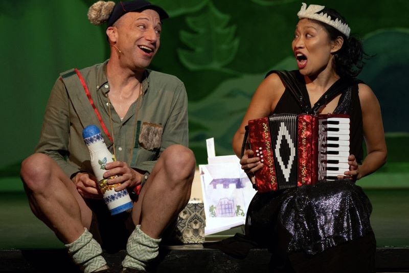 Two performers on stage in costume, smiling and singing. The man wears a hat with ears, holds a decorated stick and bottle, and has fuzzy socks; the woman wears a sparkly dress with a tiara and plays a red accordion. A hand-drawn backdrop of a house is visible between them.