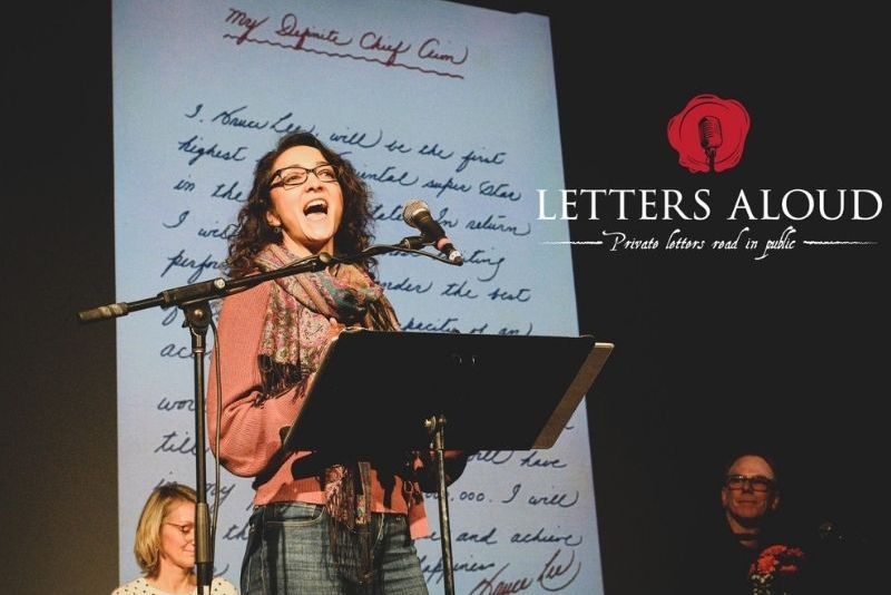 Woman standing at a microphone with a large handwritten letter behind her