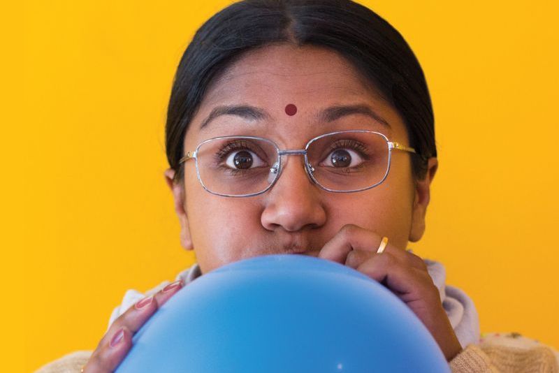 Person with glasses and a red bindi blowing up a blue balloon, against a bright yellow background.