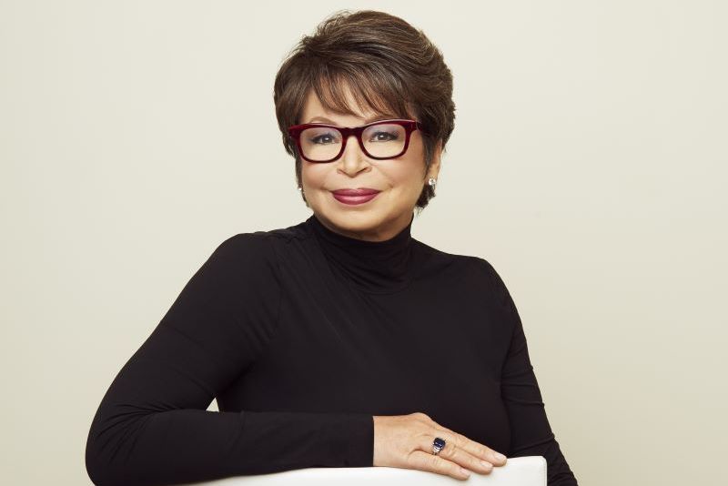 Woman wearing a black turtleneck and red-framed glasses, seated and smiling with one arm resting on a white chair, against a plain light background.