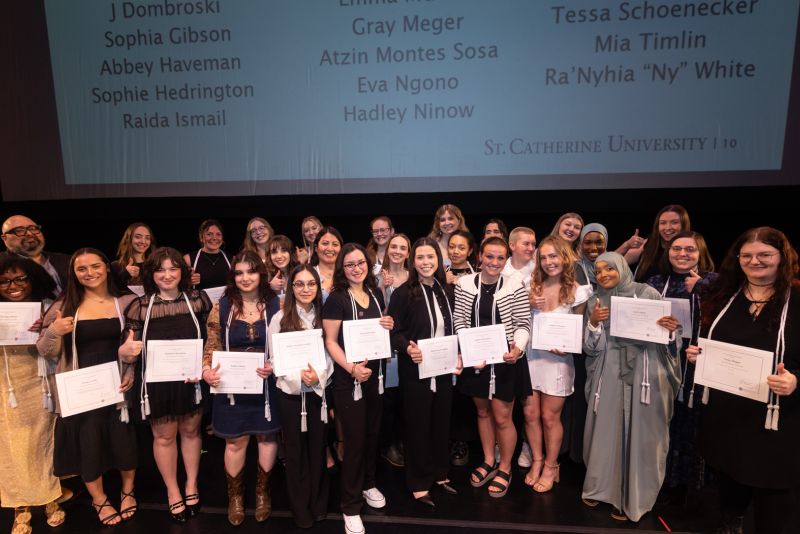 The Antonian Scholars stand on stage, smiling and holding up paper certificates.