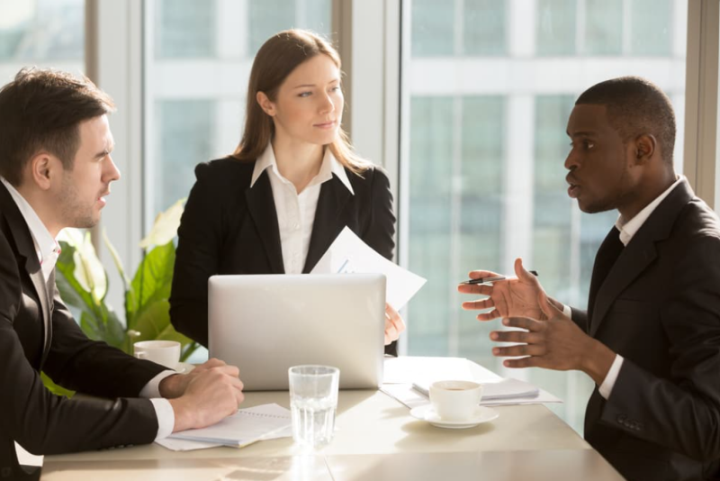 A trio of two businessmen and one businesswoman intently discuss a project in a high-rise business office. 