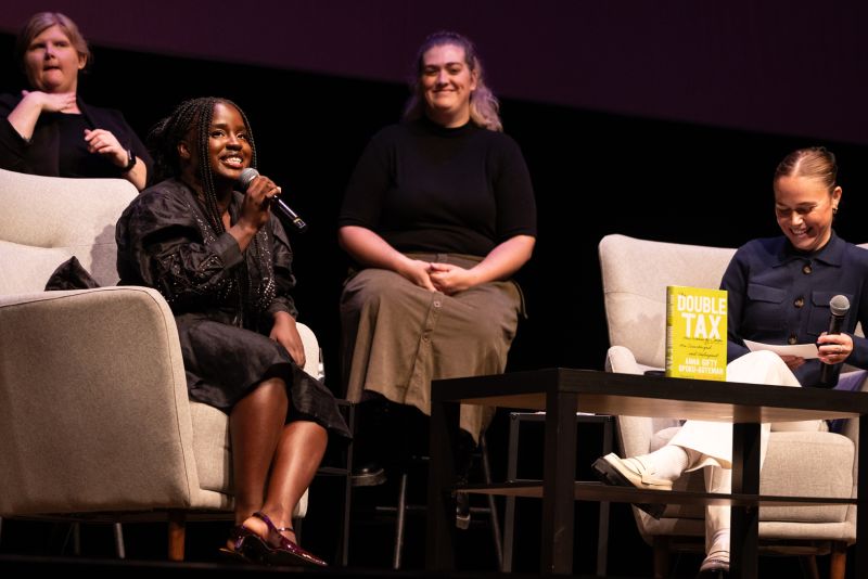 Anna Gifty Opoku-Agyeman sits on stage and speaks into a microphone. Sitting across from her is Hope Walz and behind them sit two sign language interpreters.
