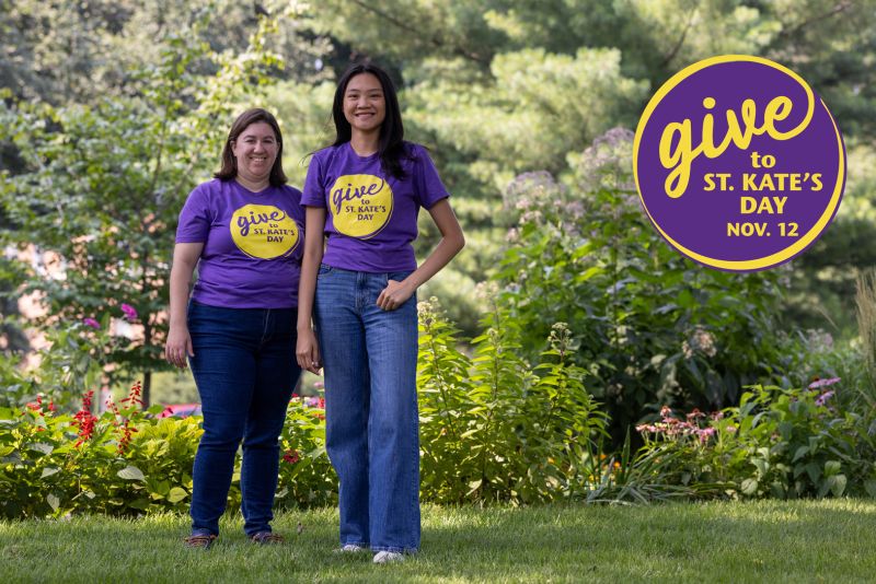 Two people wearing purple T-shirts and jeans stand in a green, leafy setting. A logo reads "Give to St. Kate's Day Nov. 12"