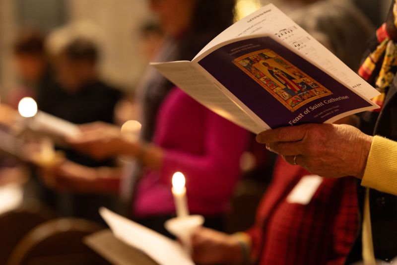 At the Feast of Saint Catherine prayer service in the Chapel, hands hold a prayer book. Lit candles can be seen in the blurred background.