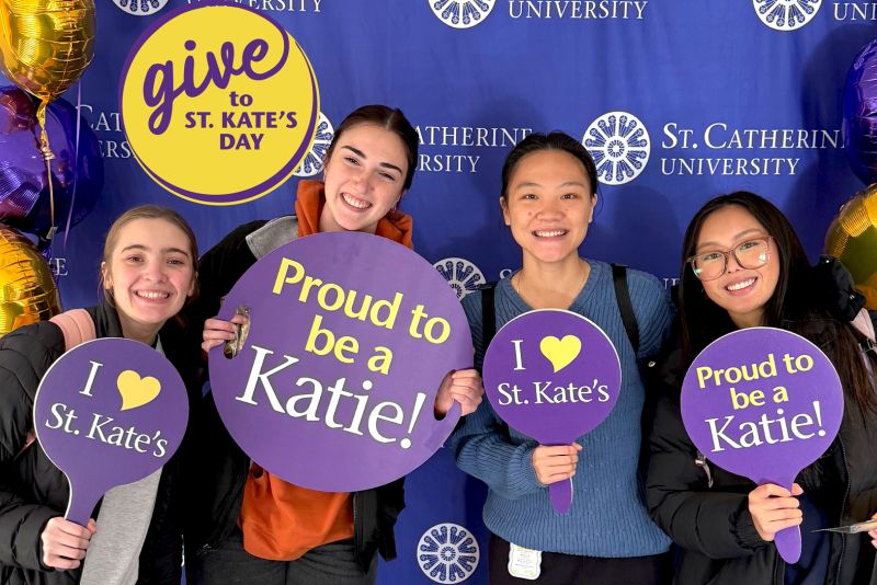 Four students pose with purple signs that say "Proud to be a Katie!" and "I heart St. Kate's"