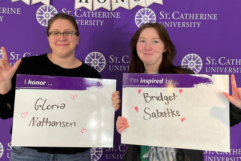 Two people standing in front of a purple backdrop each hold up whiteboards with names written on them. 
