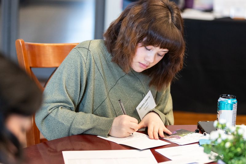 Student writing a note at a table during a campus event, with papers, pen, and beverage visible nearby.