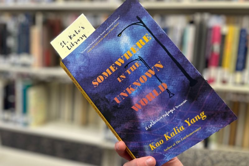 A hand holds a copy of "Somewhere in the Unknown World," with St. Kate's library shelves in background