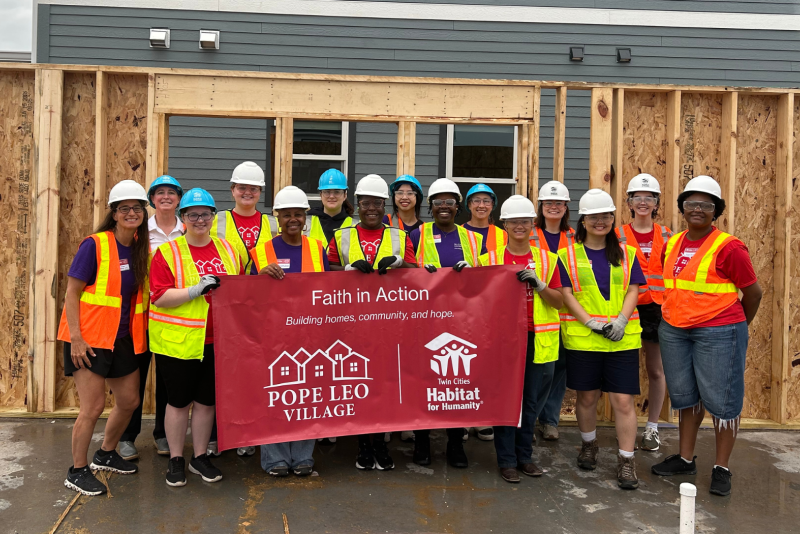 A group of people wearing neon safety vests and hard hats pose with a red banner at a construction site.
