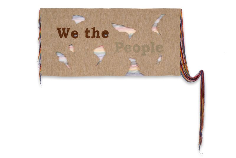 Fiber artwork on tan fabric reading “We the People,” with irregular openings revealing layers of multicolored thread. Long strands of colorful fiber extend from both sides, including a cascading bundle on the right.