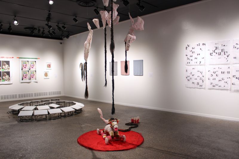 Gallery installation featuring suspended braided sculptures, textile and mixed-media wall works, a circular floor arrangement of small platforms, and a toy horse sculpture displayed on a red rug.
