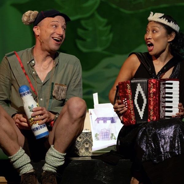 Two performers on stage in costume, smiling and singing. The man wears a hat with ears, holds a decorated stick and bottle, and has fuzzy socks; the woman wears a sparkly dress with a tiara and plays a red accordion. A hand-drawn backdrop of a house is visible between them.