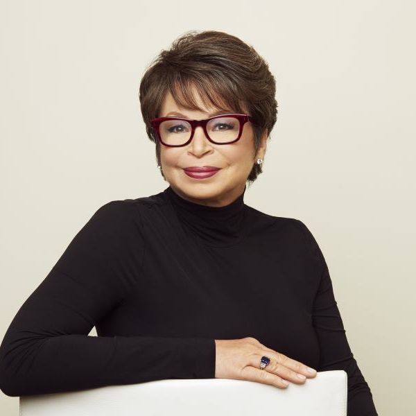 Woman wearing a black turtleneck and red-framed glasses, seated and smiling with one arm resting on a white chair, against a plain light background.