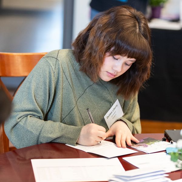 Student writing a note at a table during a campus event, with papers, pen, and beverage visible nearby.