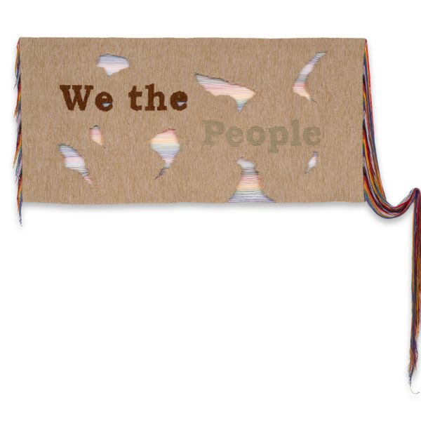 Fiber artwork on tan fabric reading “We the People,” with irregular openings revealing layers of multicolored thread. Long strands of colorful fiber extend from both sides, including a cascading bundle on the right.